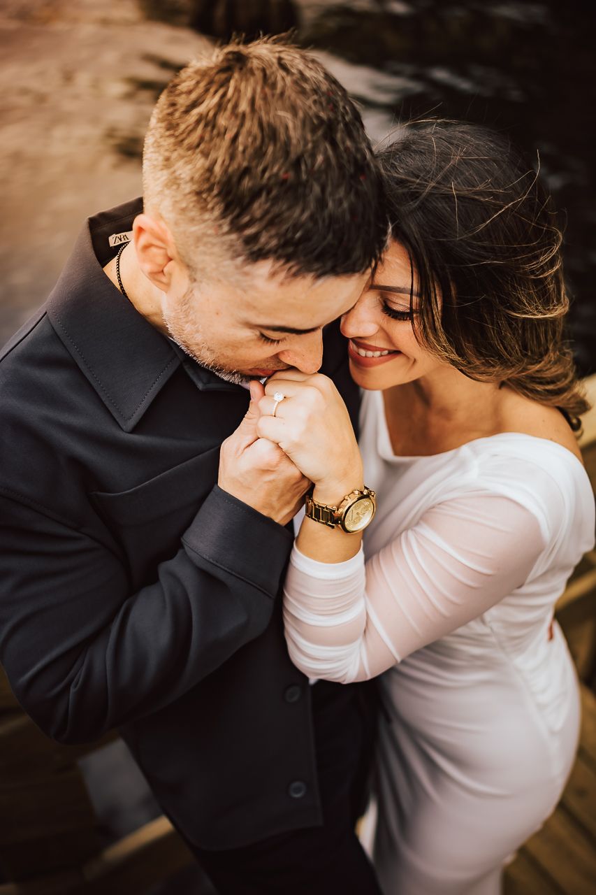 Romantic moment between couple in formal attire with man in black suit kissing woman's hand during golden hour photography.