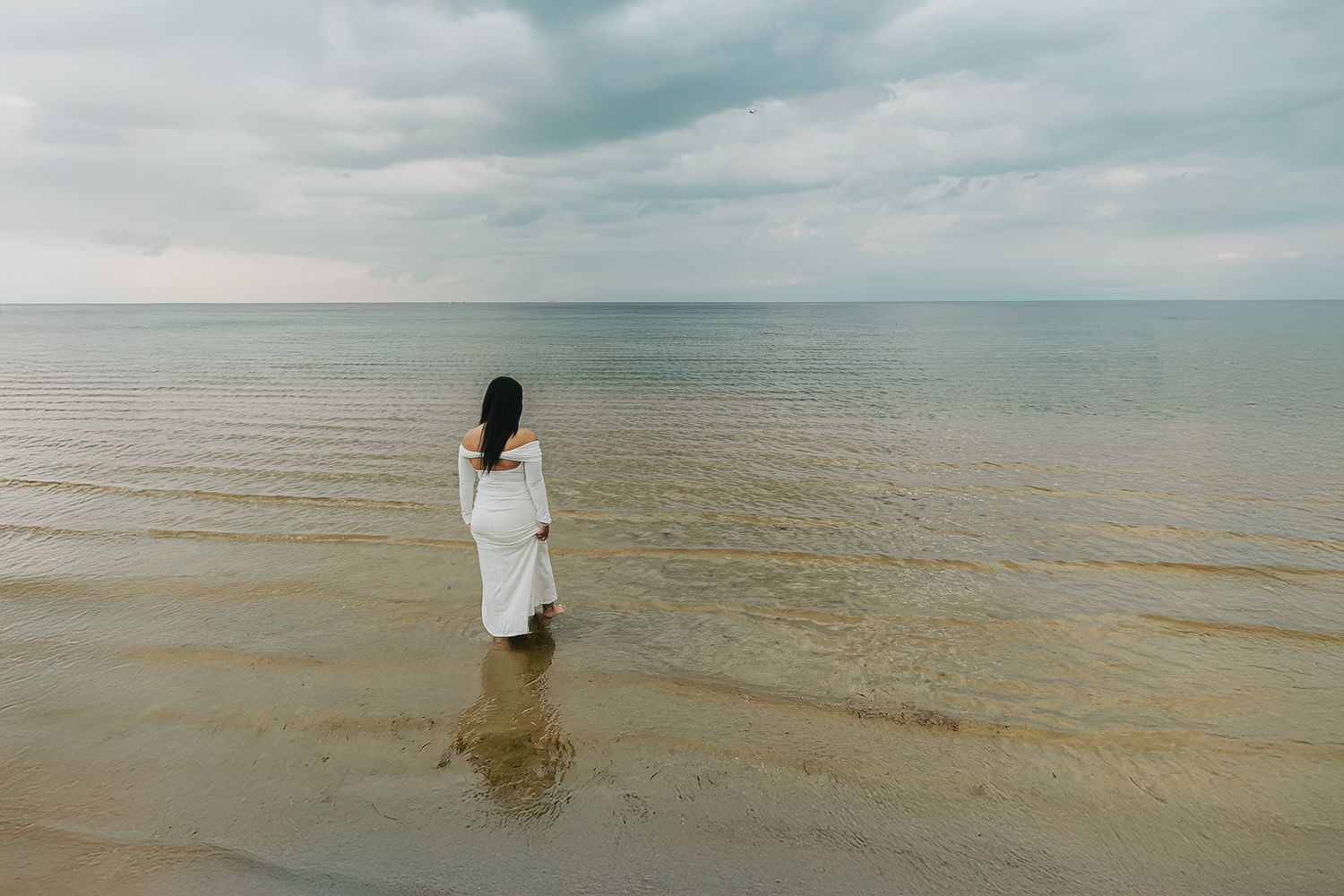 Person in white dress stands alone at the edge of calm ocean waters under moody overcast sky.