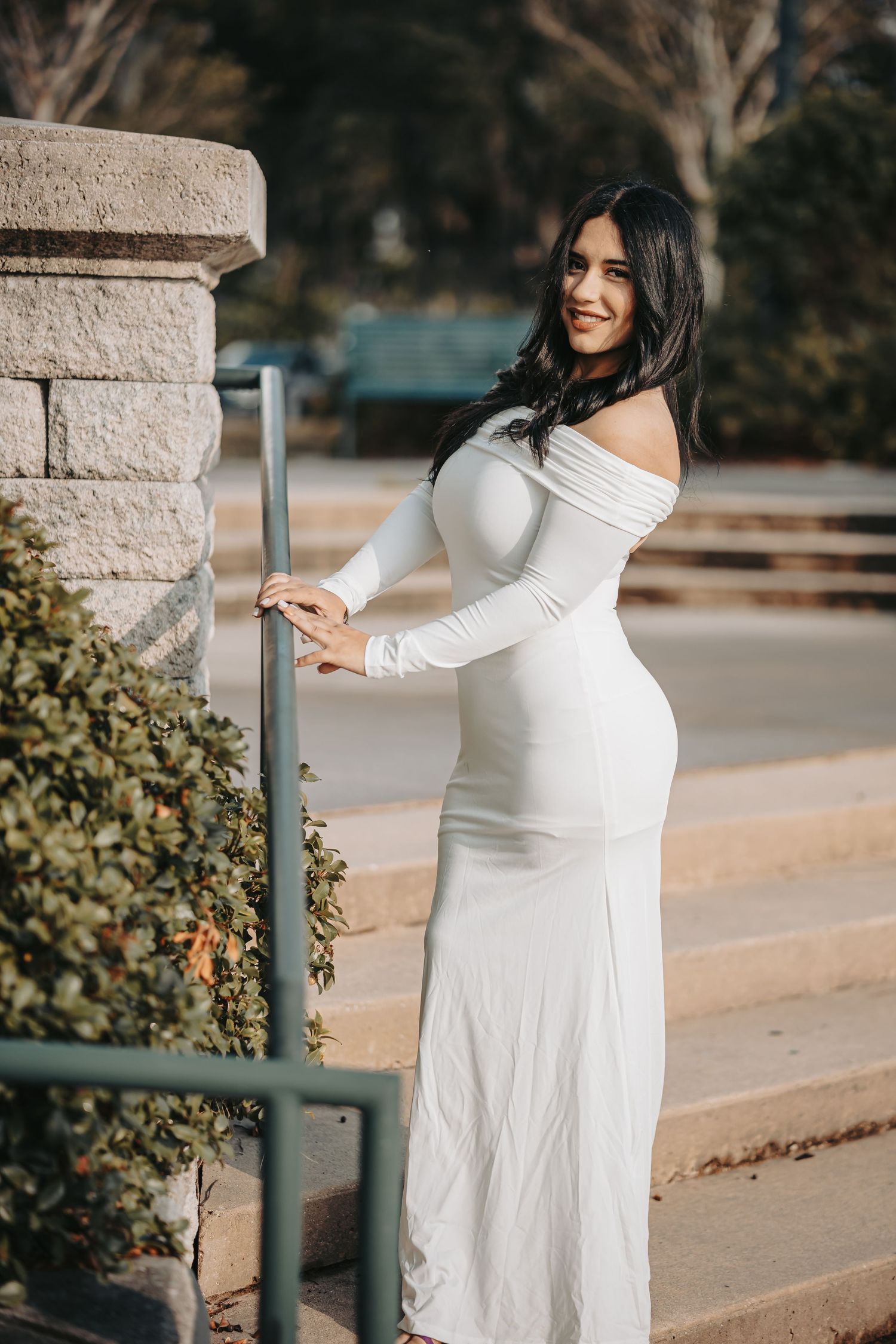 Person in elegant white off-shoulder long dress poses by a stone pillar in an outdoor garden setting.
