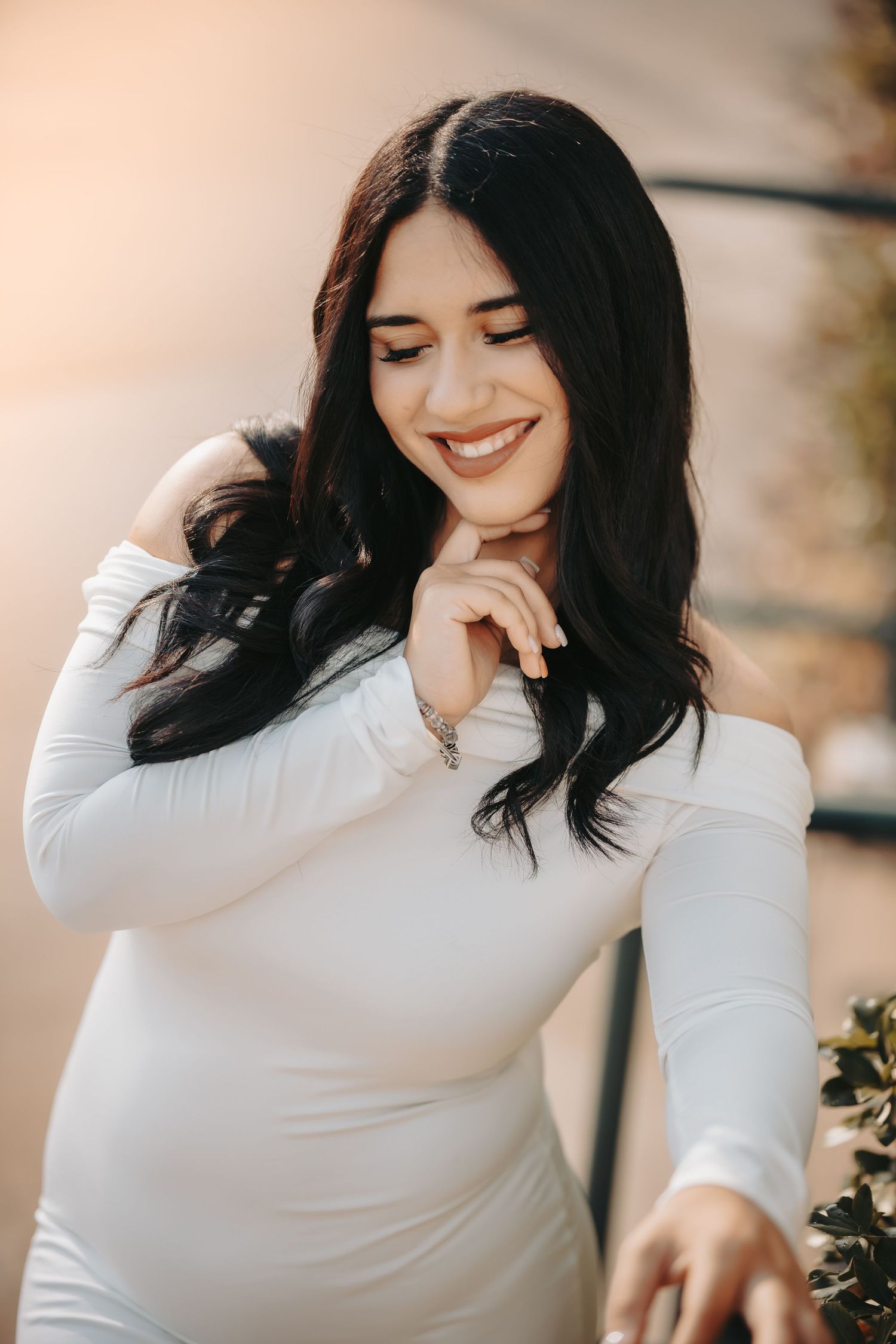 A person with dark hair smiles warmly while wearing a fitted white long-sleeve top in soft natural lighting.