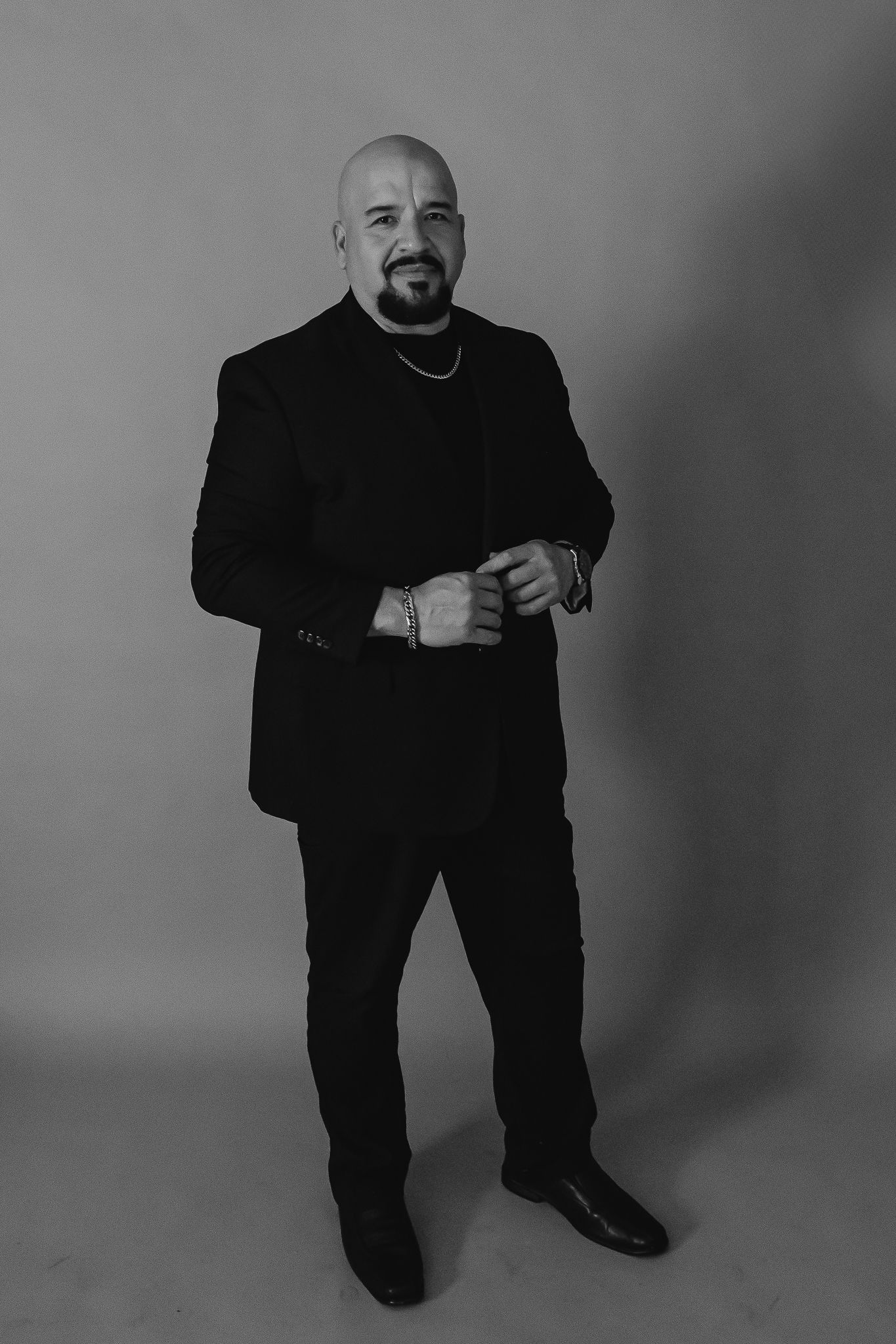 Man in all black formal attire poses against grey backdrop in studio portrait.