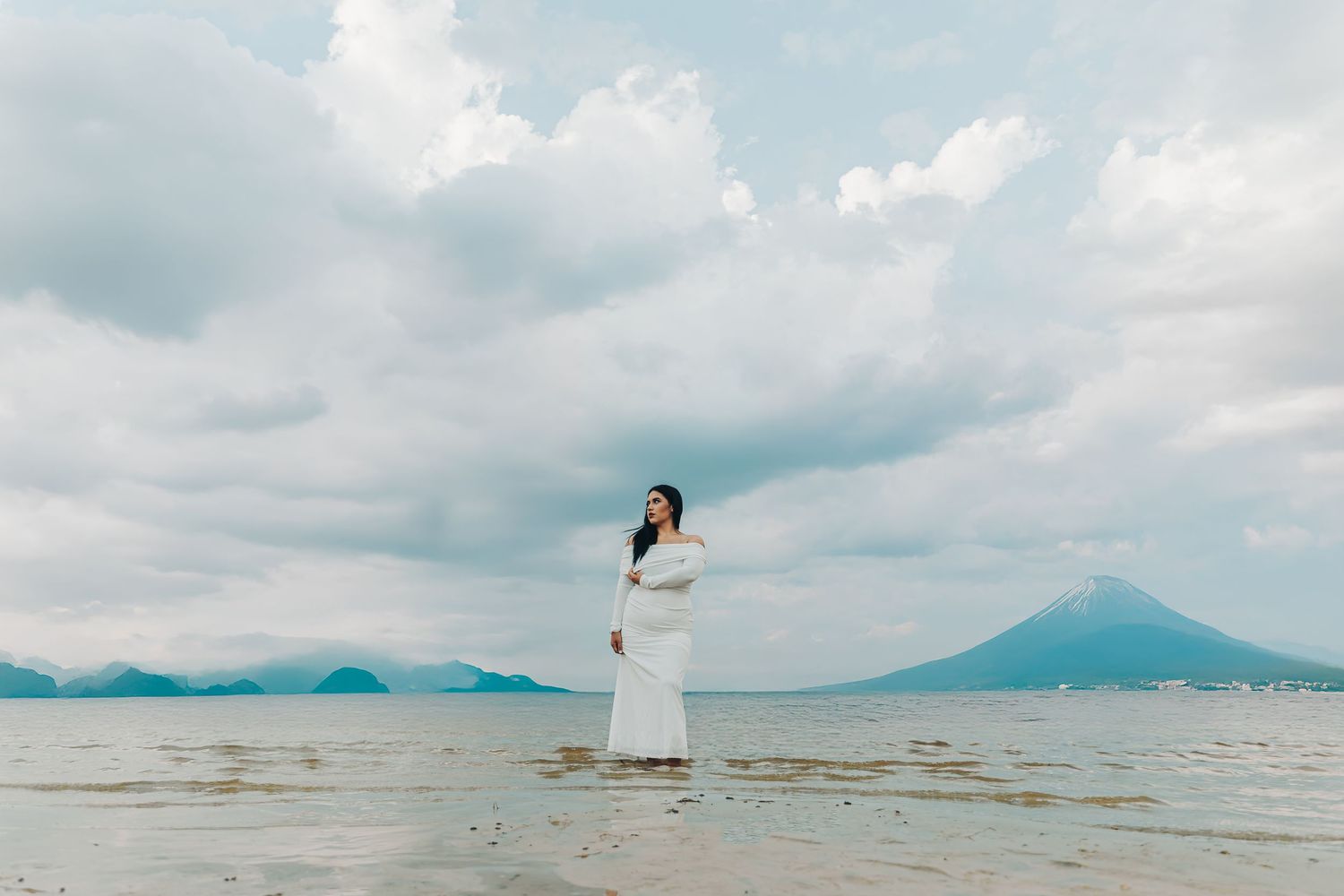 Figure in white dress stands in shallow water with dramatic mountain landscape and cloudy sky in the background.