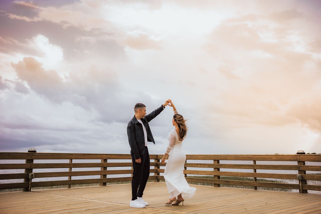 A couple dances together on a wooden deck overlooking a scenic cloudy sunset sky.