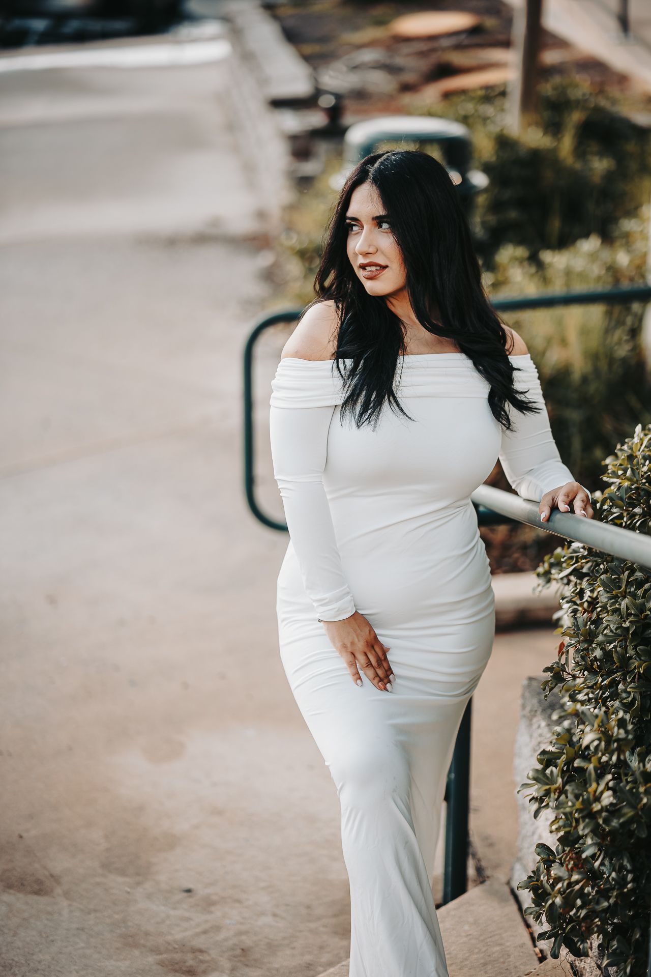 Long-sleeved off-shoulder white dress worn outdoors on a sunny day with garden path and greenery in background.