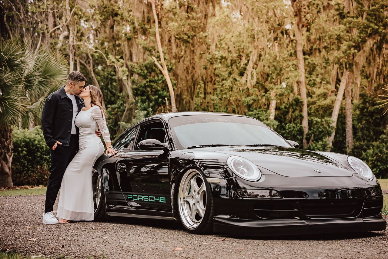 Newlyweds pose with sleek black Porsche GT3 sports car in a wooded outdoor setting.