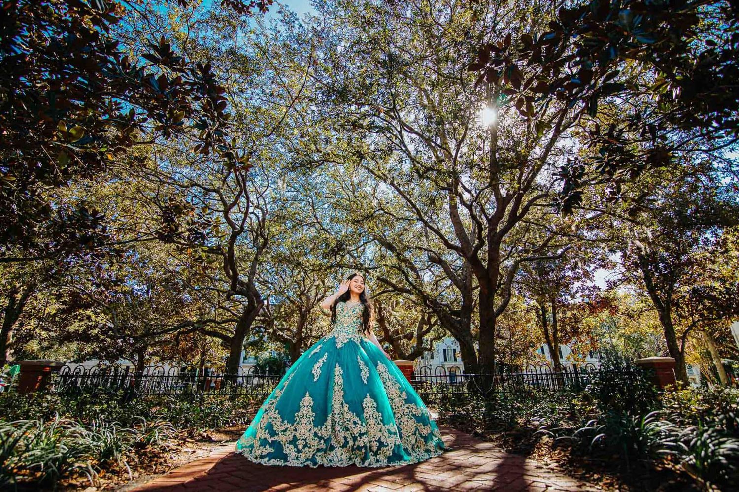 Beautiful turquoise quinceañera gown with floral details photographed in a sunlit garden path surrounded by trees.