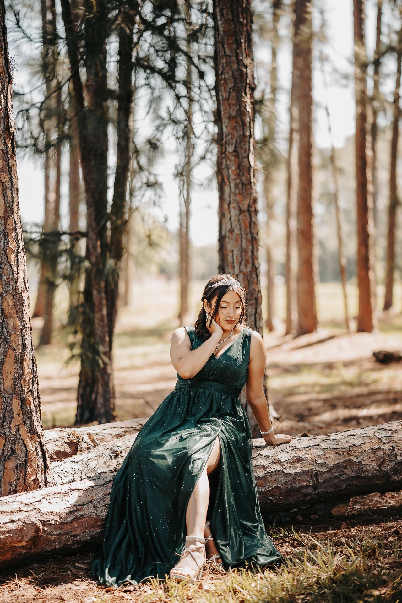 Person in emerald green formal dress poses elegantly on fallen log in sunlit forest.