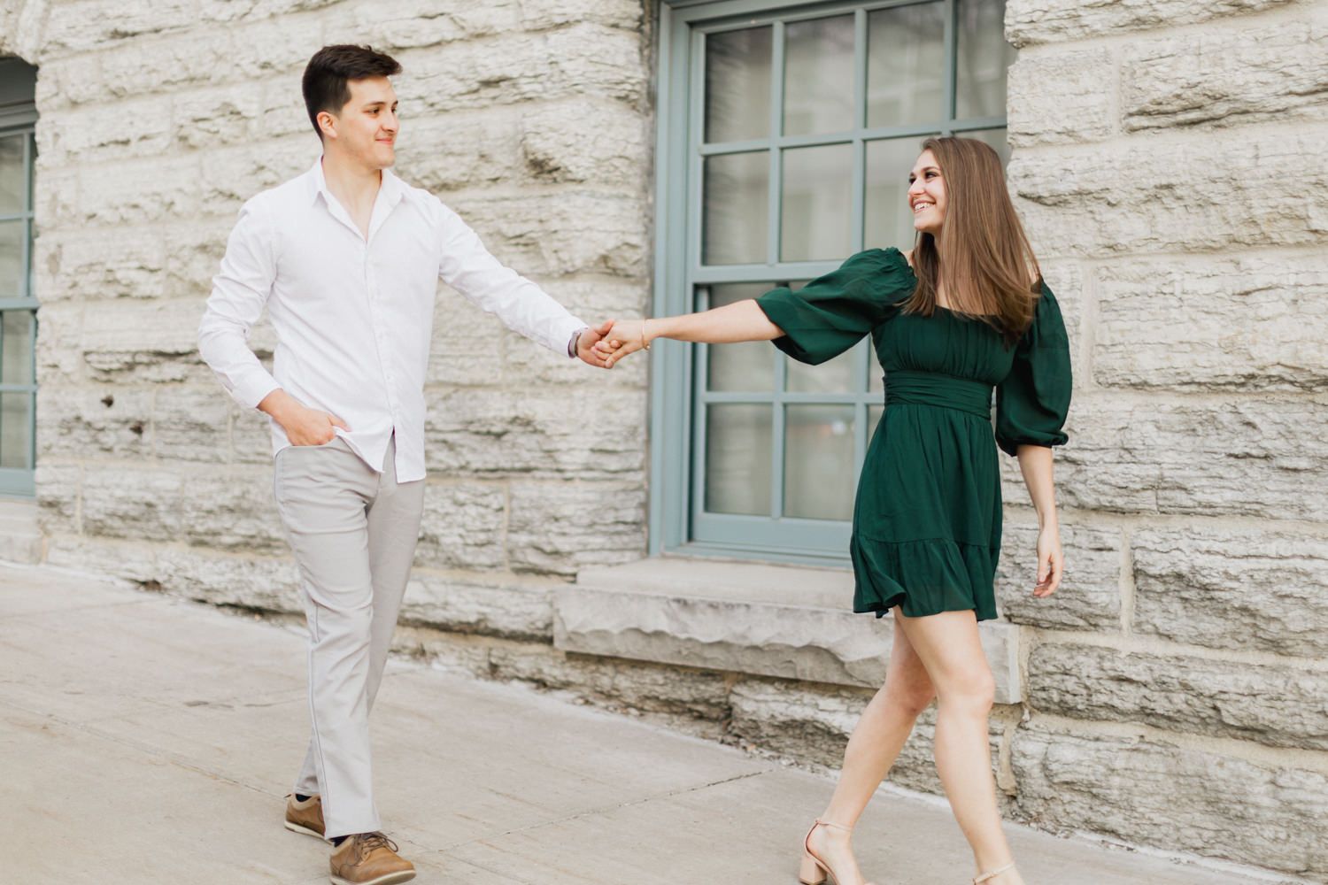 Couple holds hands while dancing outside stone building.