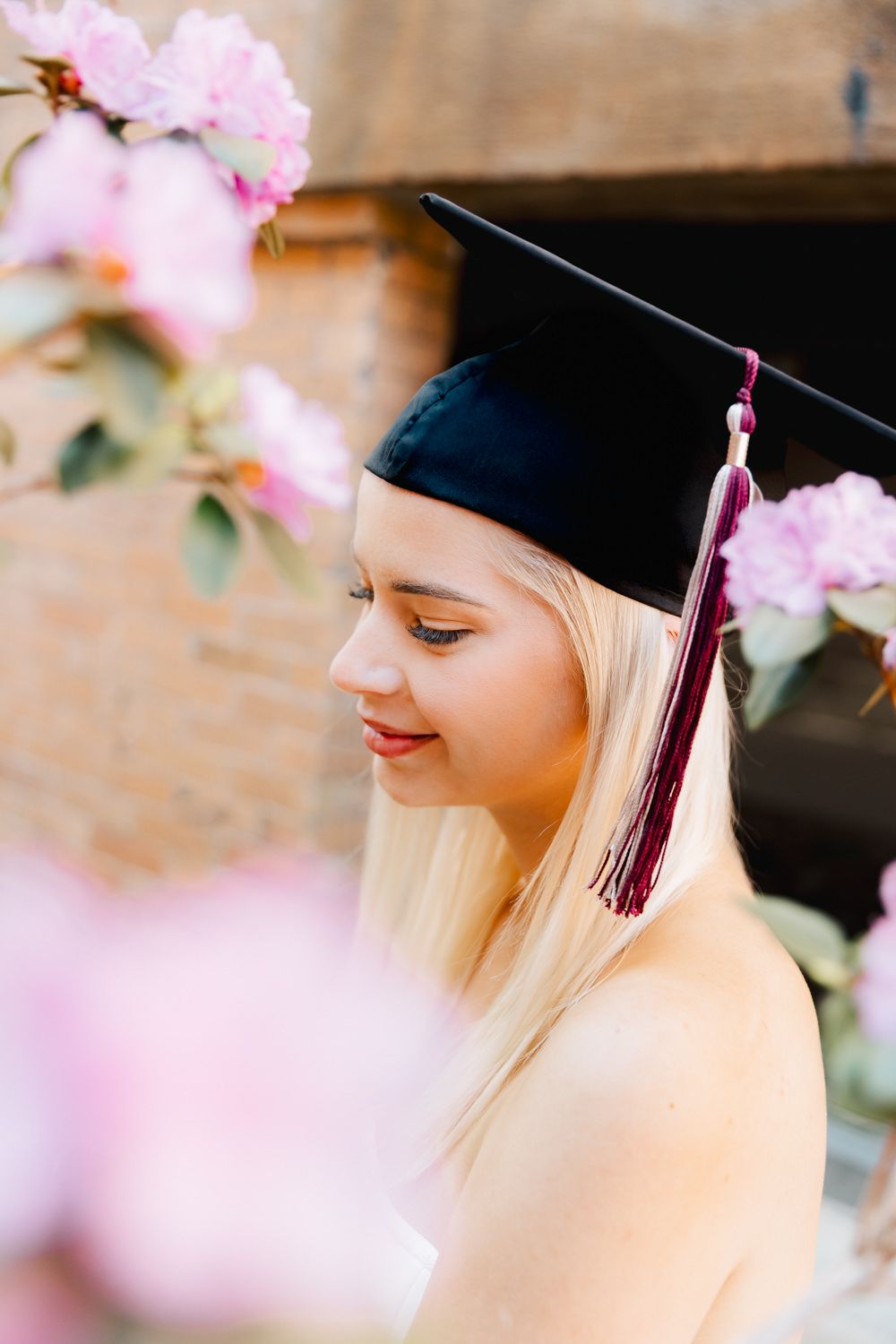 Graduate wearing black cap and tassel smiles while standing among pink cherry blossoms.