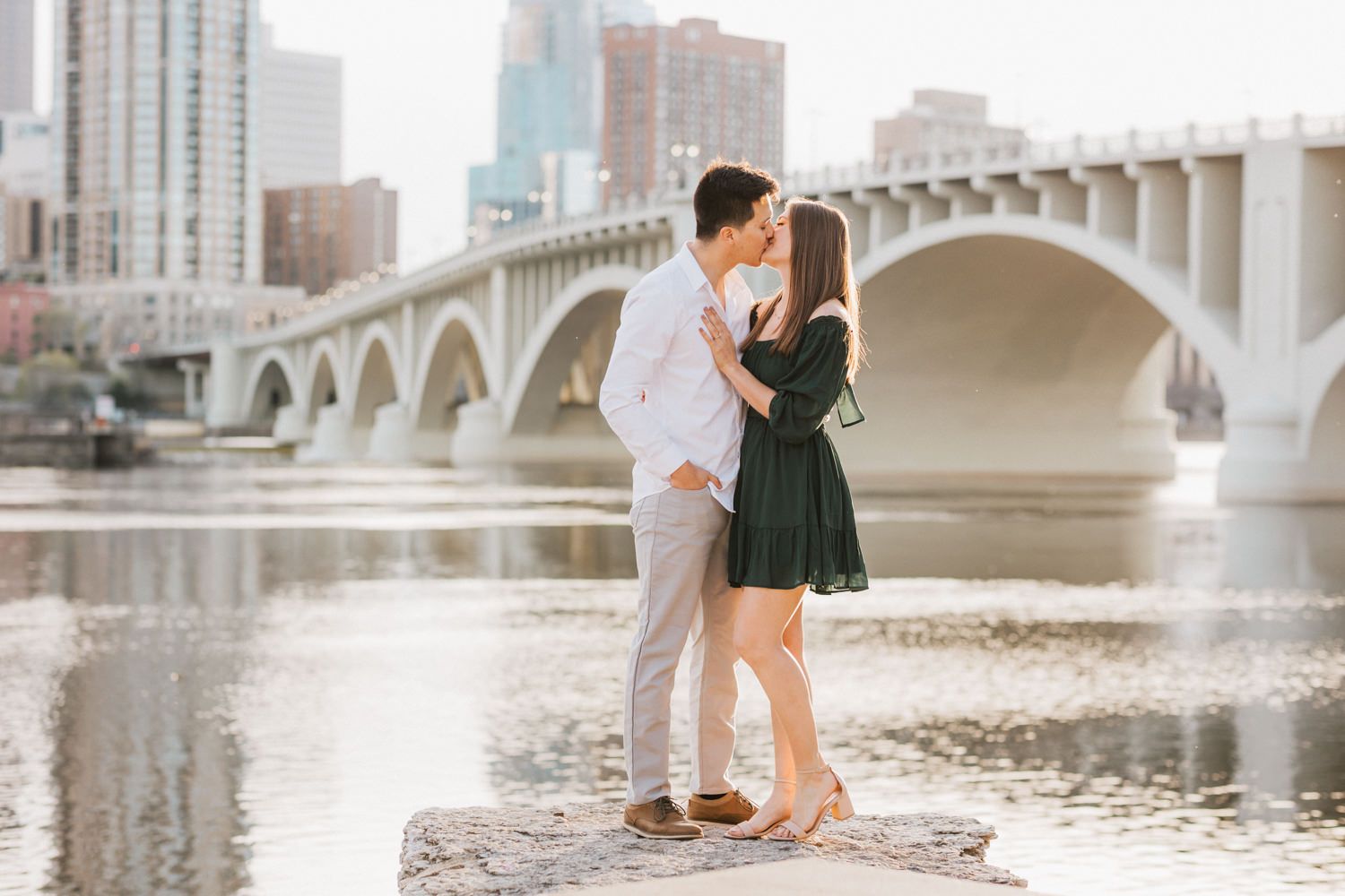 Two people in romantic poses share intimate moments along a waterfront with bridge and city skyline views during sunset.