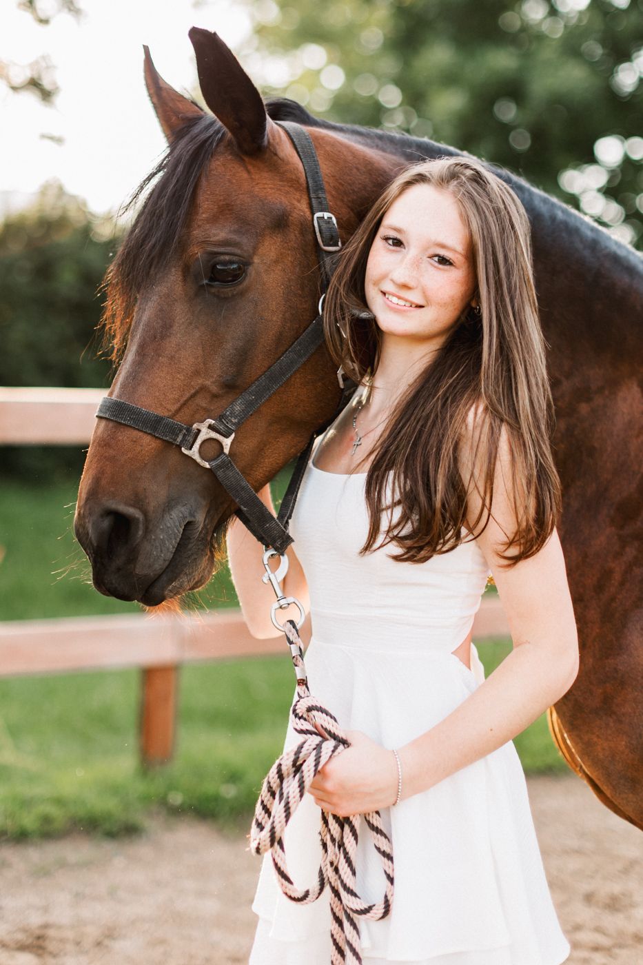 A beautiful bay horse with bridle poses with a white-dressed figure against a natural outdoor background.