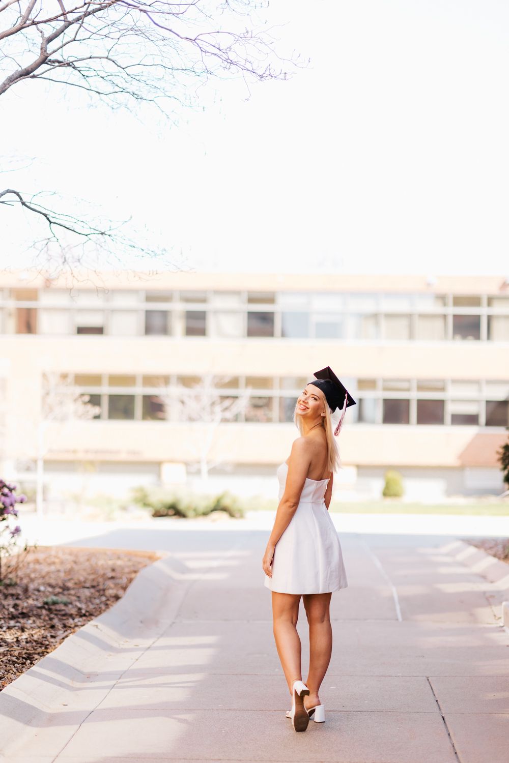 Graduate in white dress and cap walks along campus pathway.