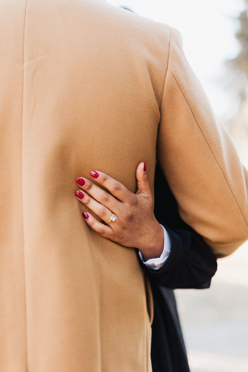 A close-up of a hand with red nails and engagement ring against a tan coat.