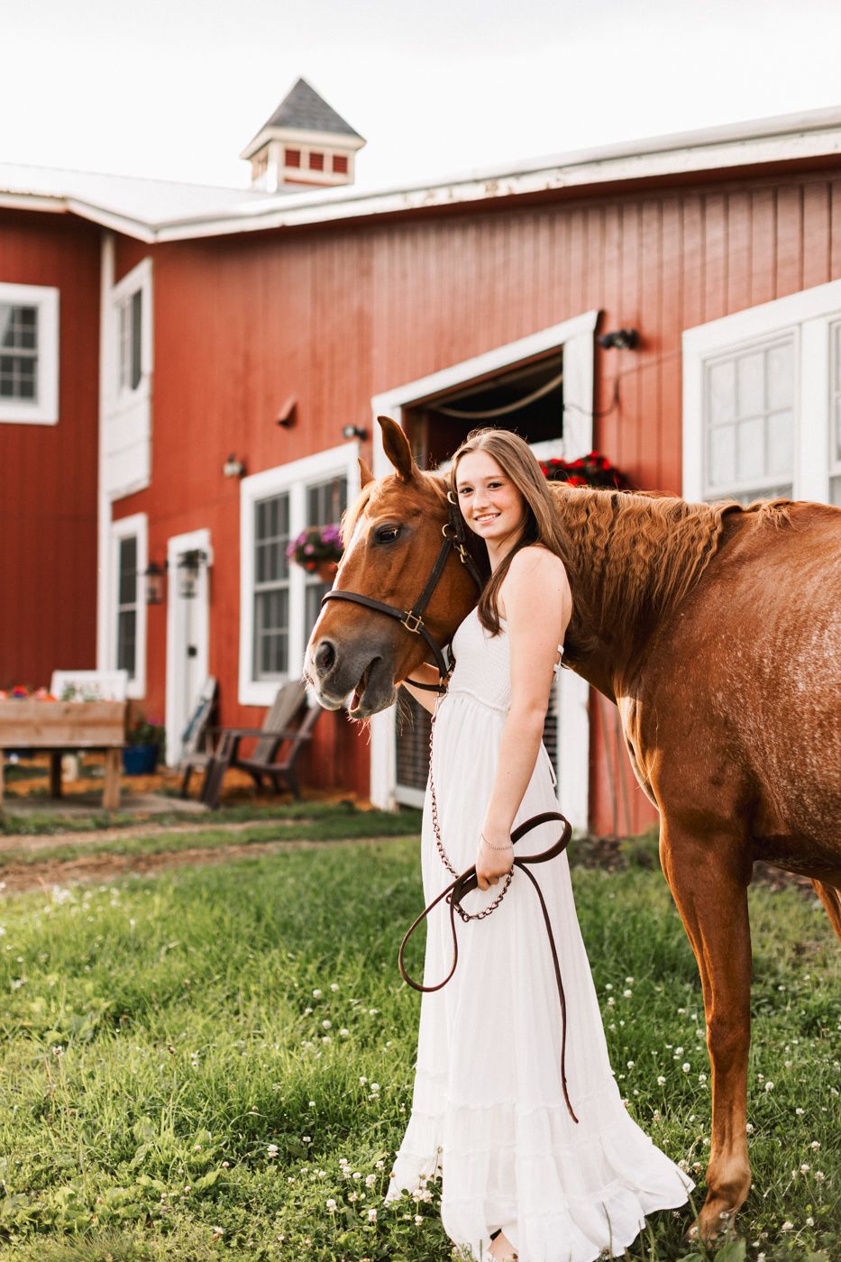 A chestnut horse and white-dressed figure pose in front of a classic red barn with white trim.