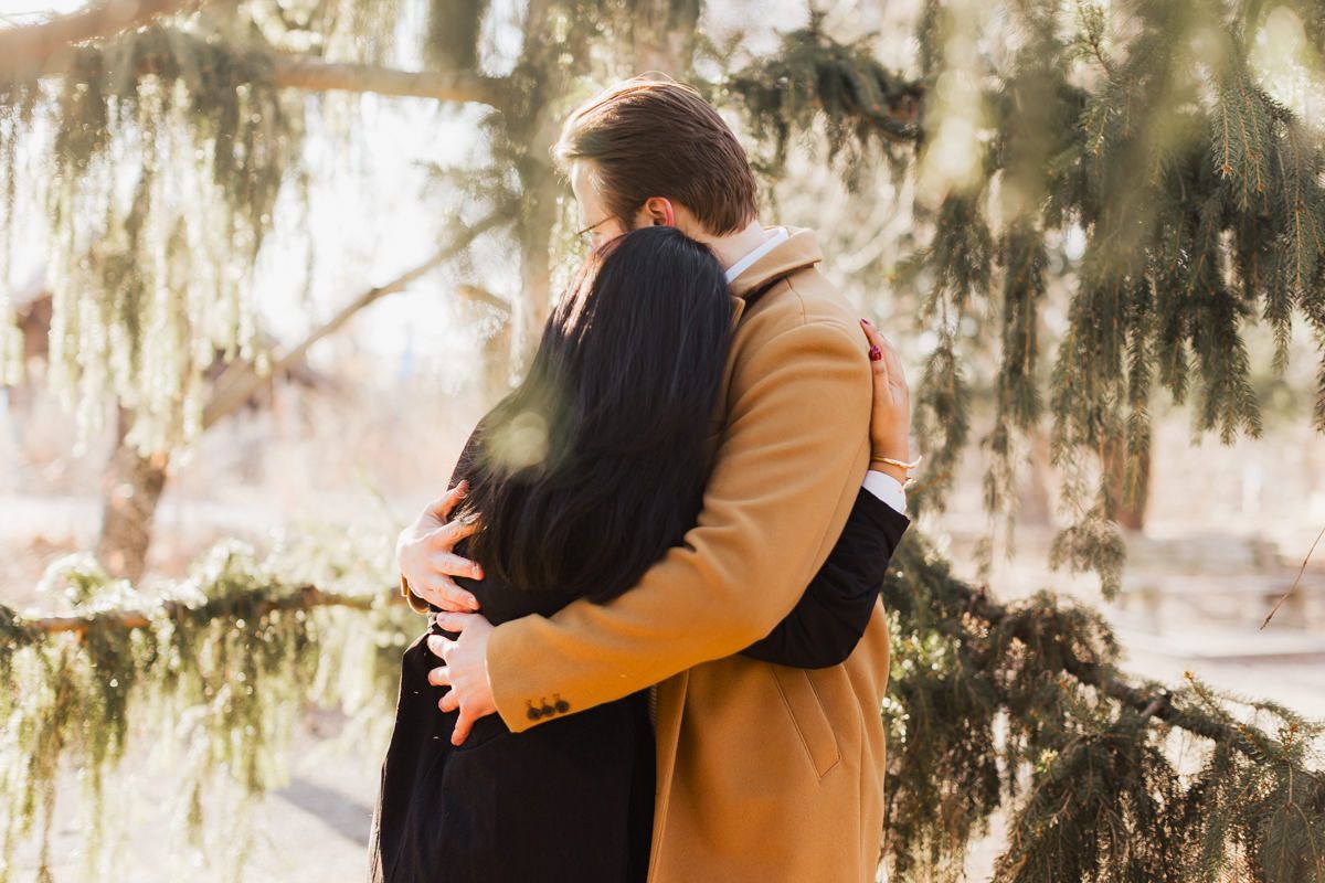 Couple in winter coats sharing an intimate embrace beneath moss-draped trees in soft sunlight.