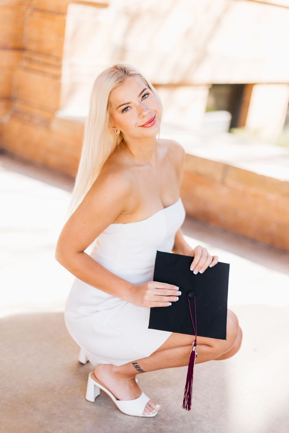 Graduate in white dress crouching with diploma holder during graduation photoshoot.