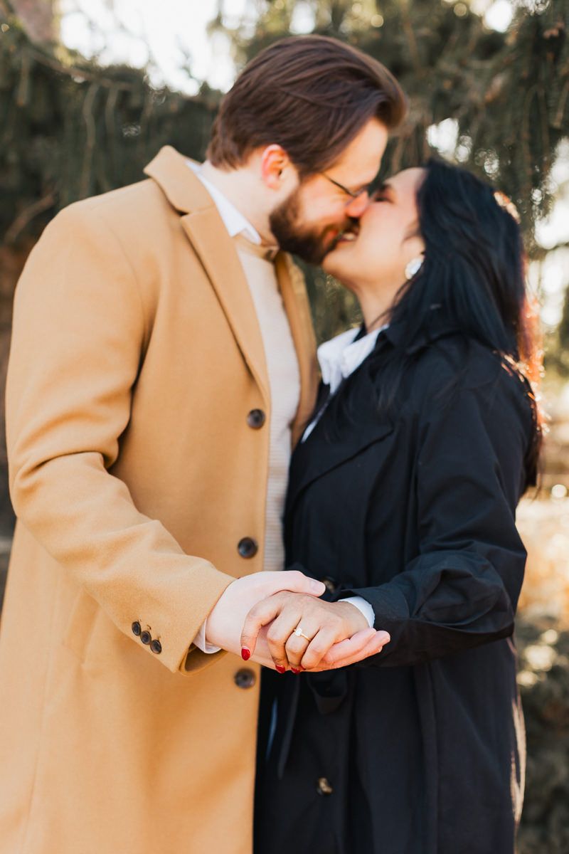 A couple shares an intimate kiss while holding hands, dressed in contrasting black and camel coats.