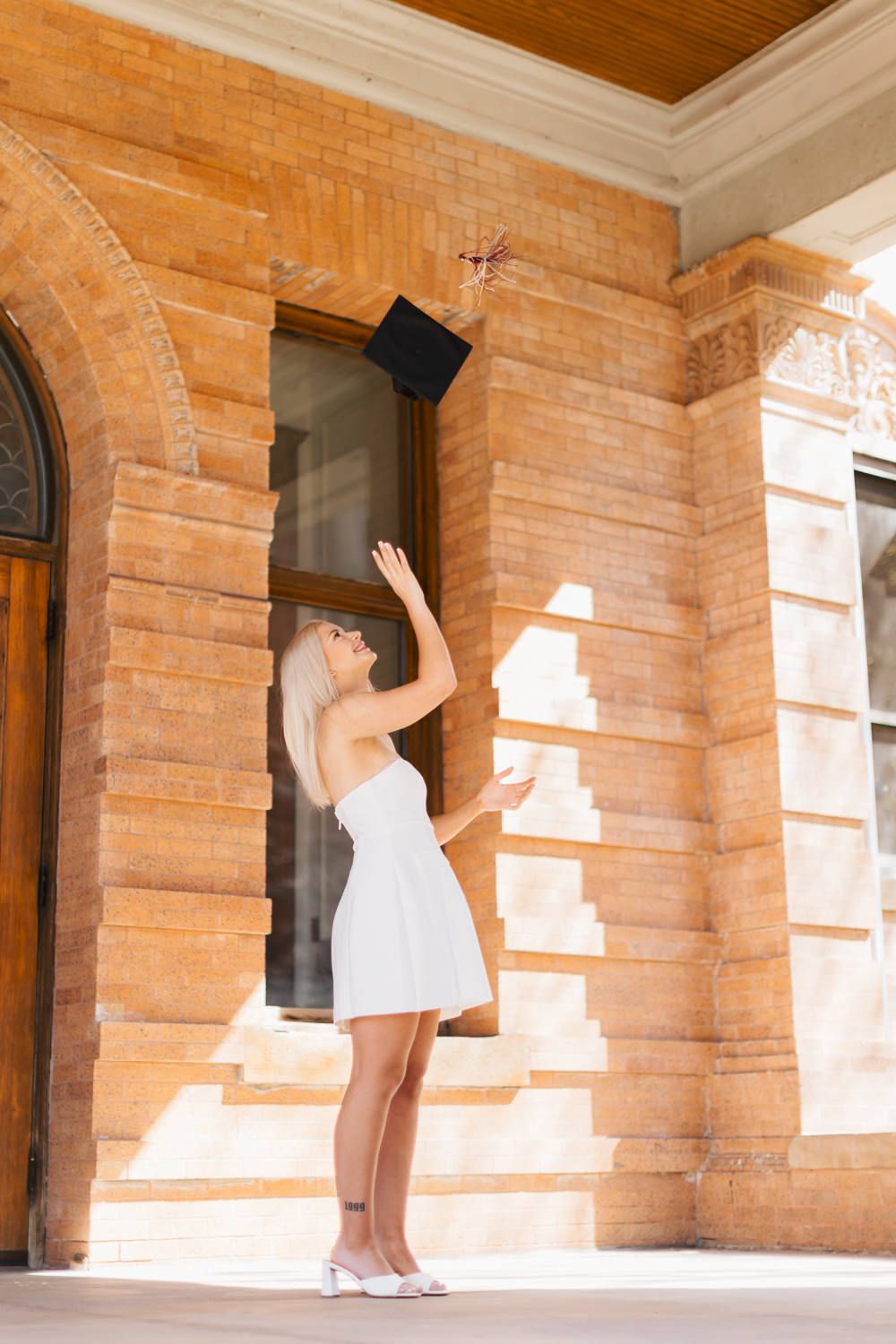 Graduate tossing graduation cap in air while wearing white dress.