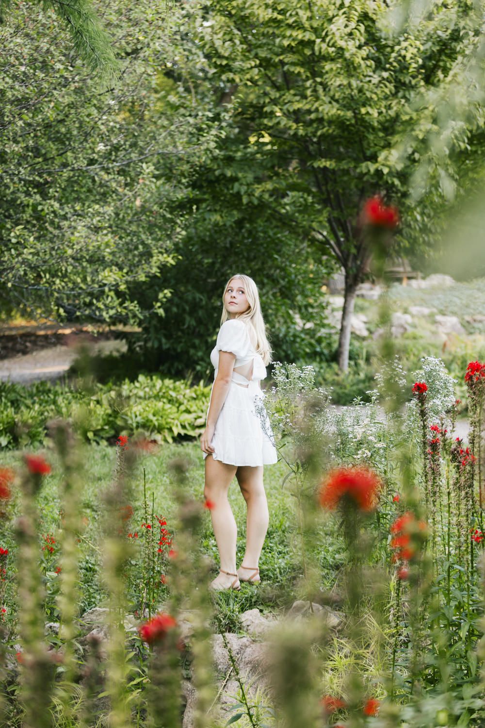 Person in white dress standing among red flowers in a lush garden setting.