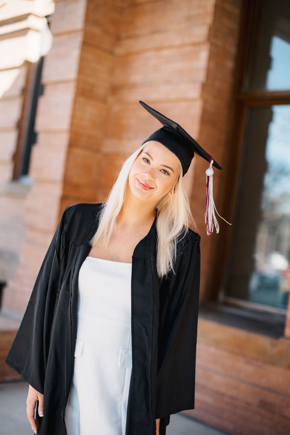 Graduate in black cap and gown smiling in front of brick building on graduation day.