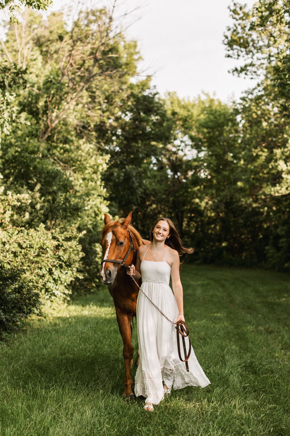 White-dressed figure stands with a chestnut horse in a sunlit meadow bordered by lush green trees.
