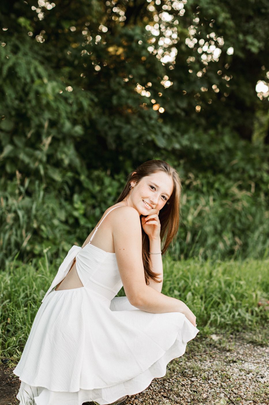 Figure in white dress poses gracefully on gravel path with bokeh-lit foliage background.