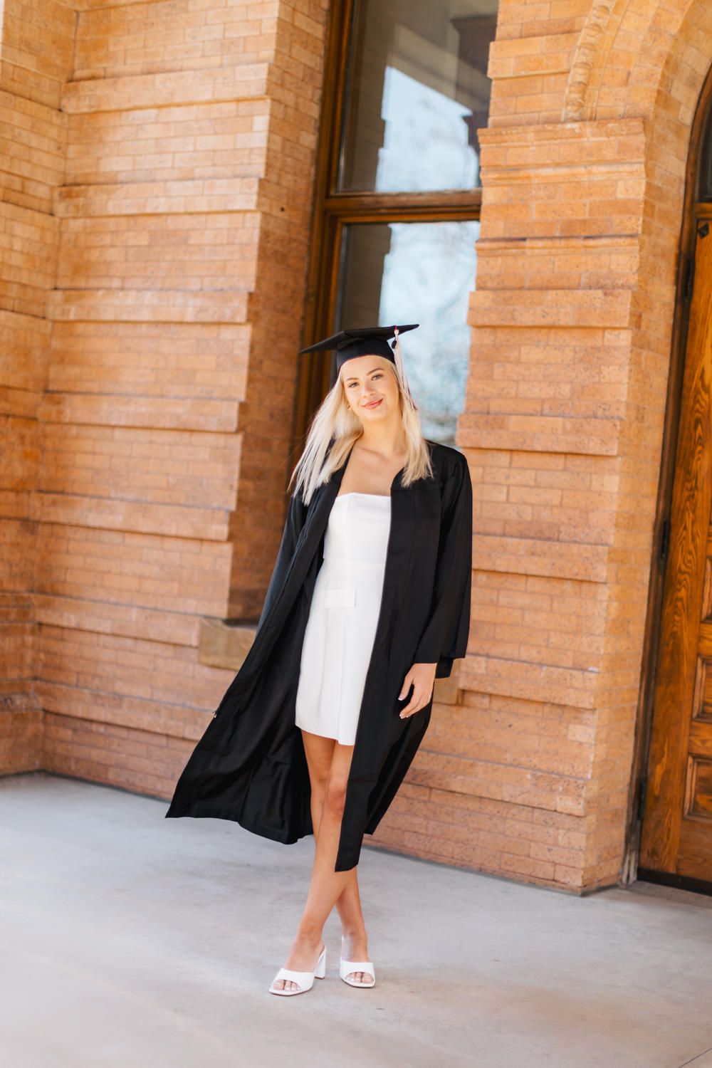 Graduate in traditional black academic regalia poses in front of brick building entrance.
