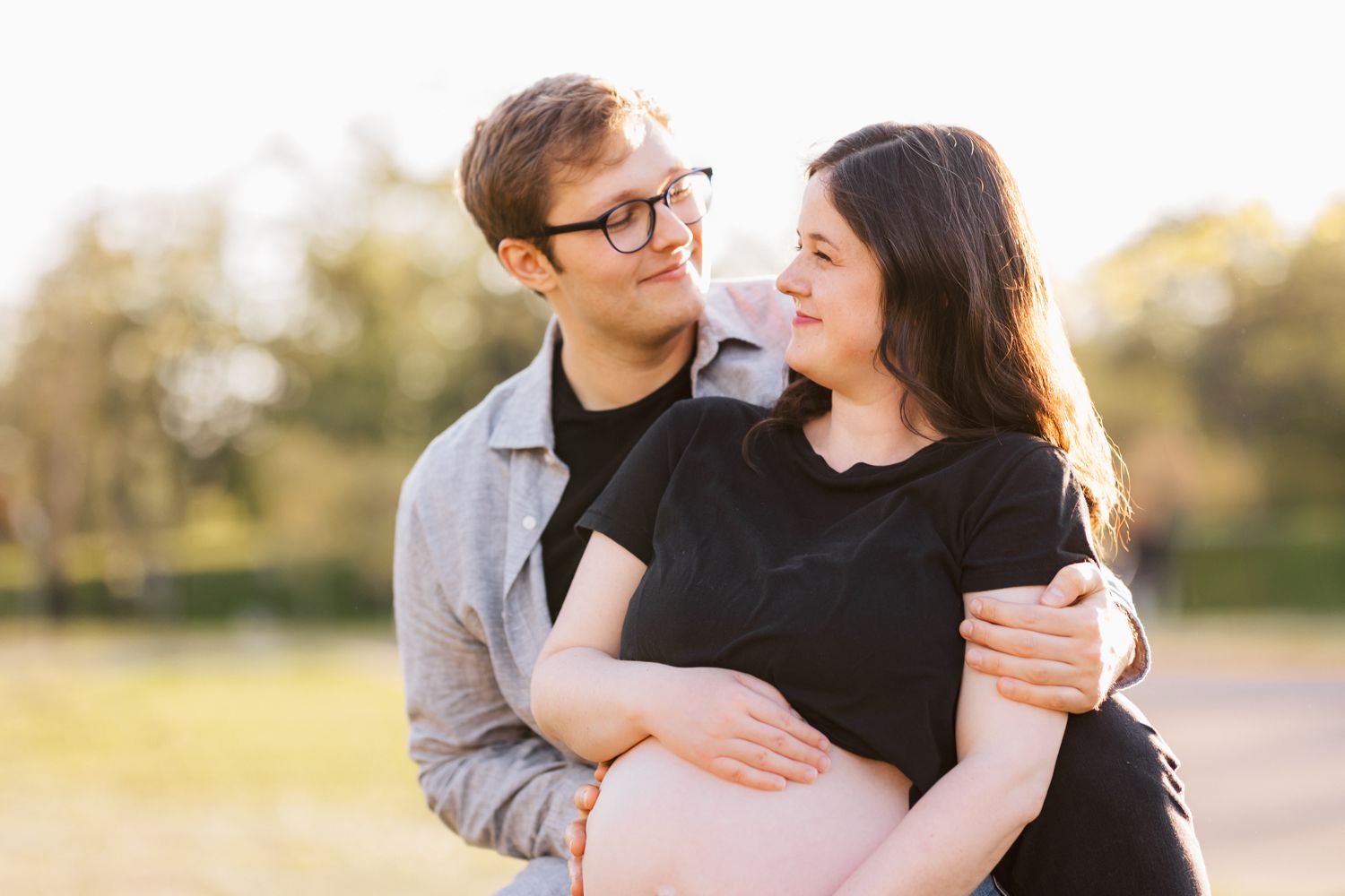 A young couple embraces outdoors during a maternity photo session at sunset in a natural setting.