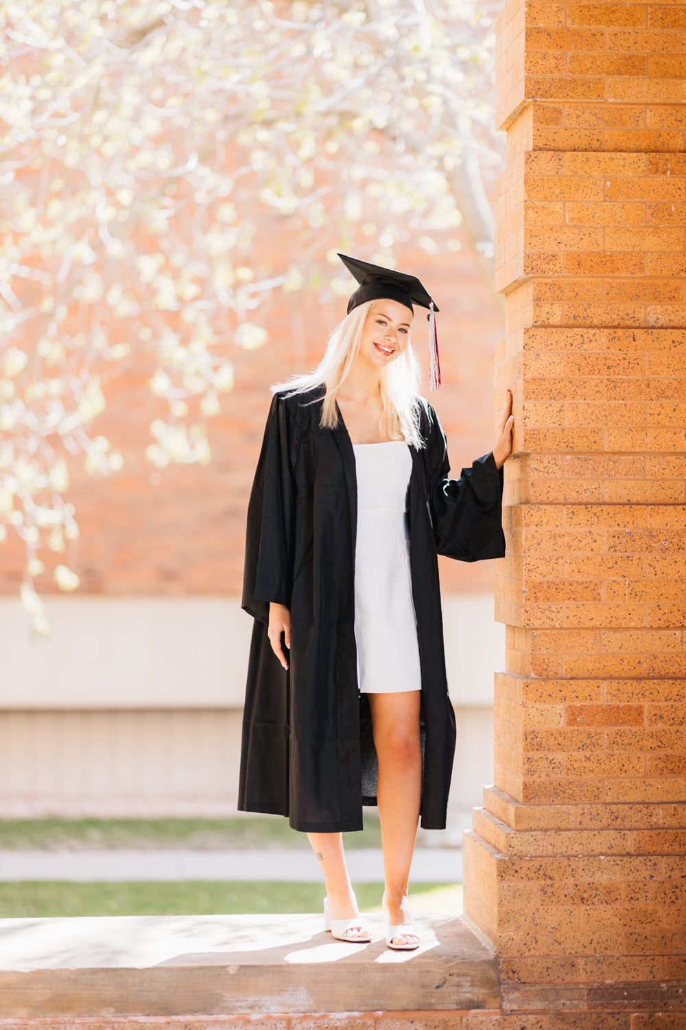 Graduate in cap and gown poses near blossoming trees and brick architecture.