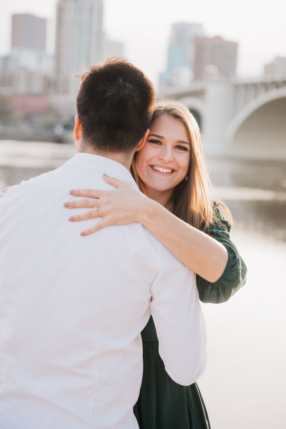 Two people embracing in a heartfelt moment by a riverfront with city buildings in the background.