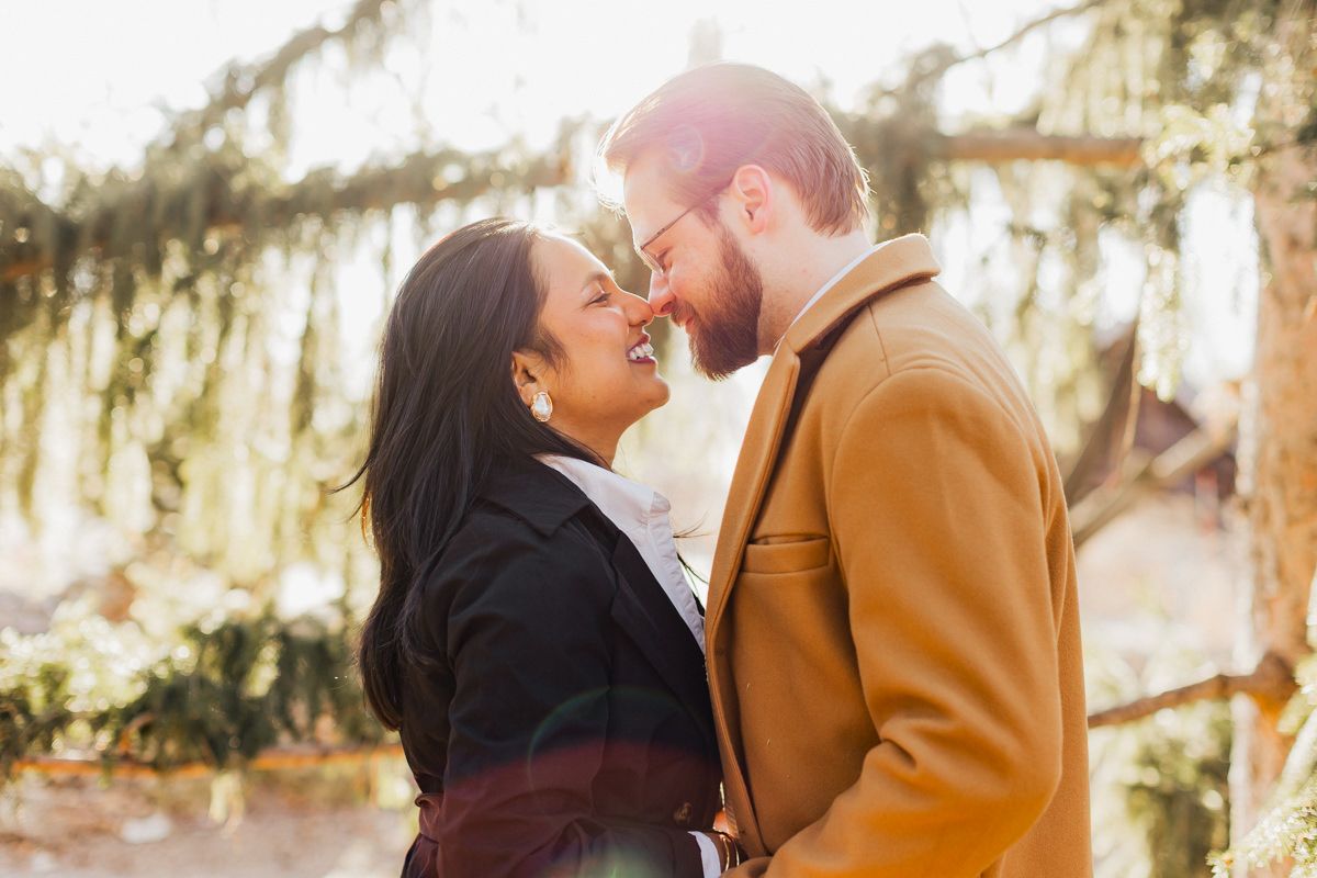 Intimate moment between couple in black and camel coats under sunlit trees.
