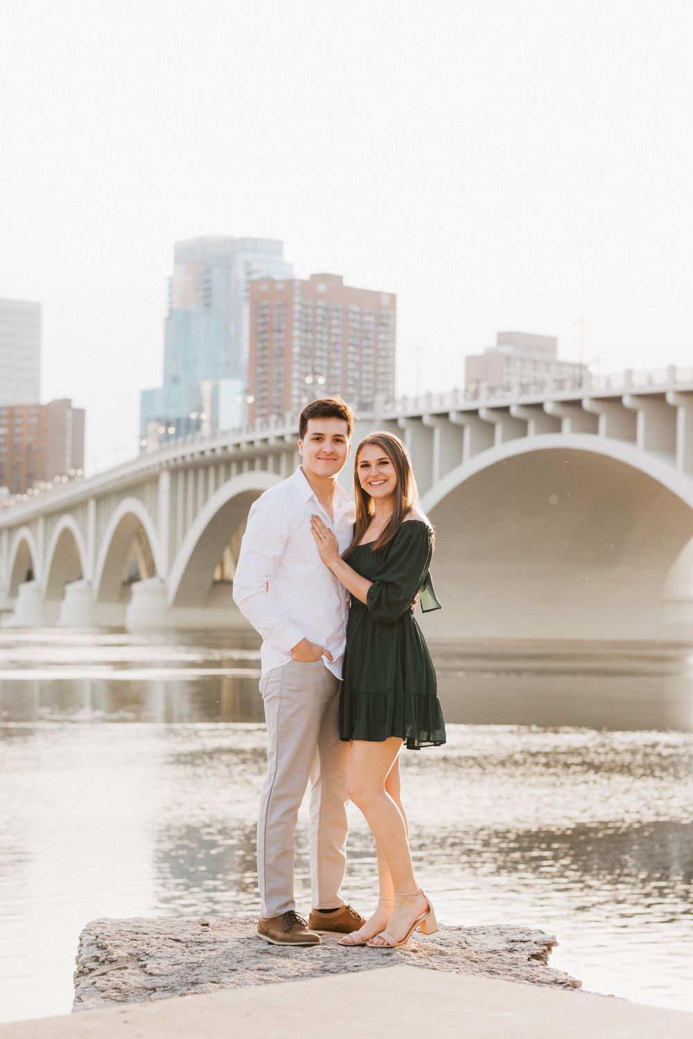 Couple in formal attire posing by riverside with arched bridge in background.