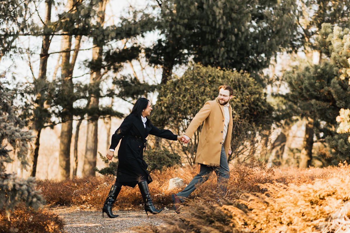 Couple in winter coats holding hands while walking through a wooded path at sunset.