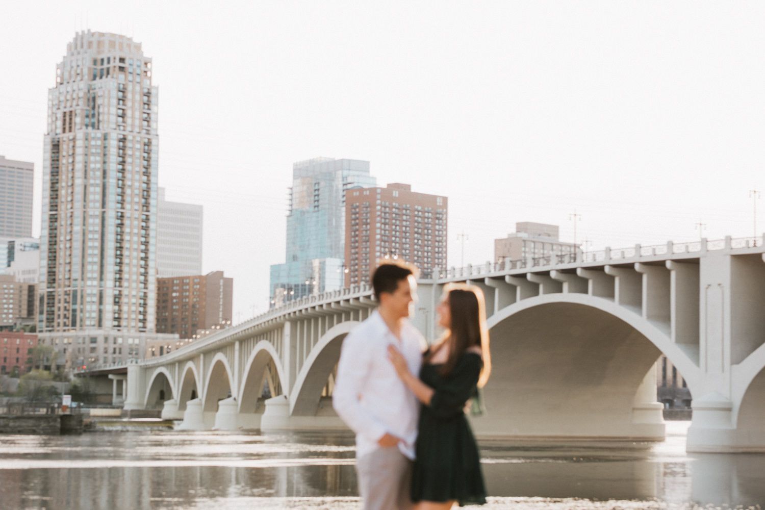 Romantic engagement photoshoot with city skyline and arched bridge in background during sunset.