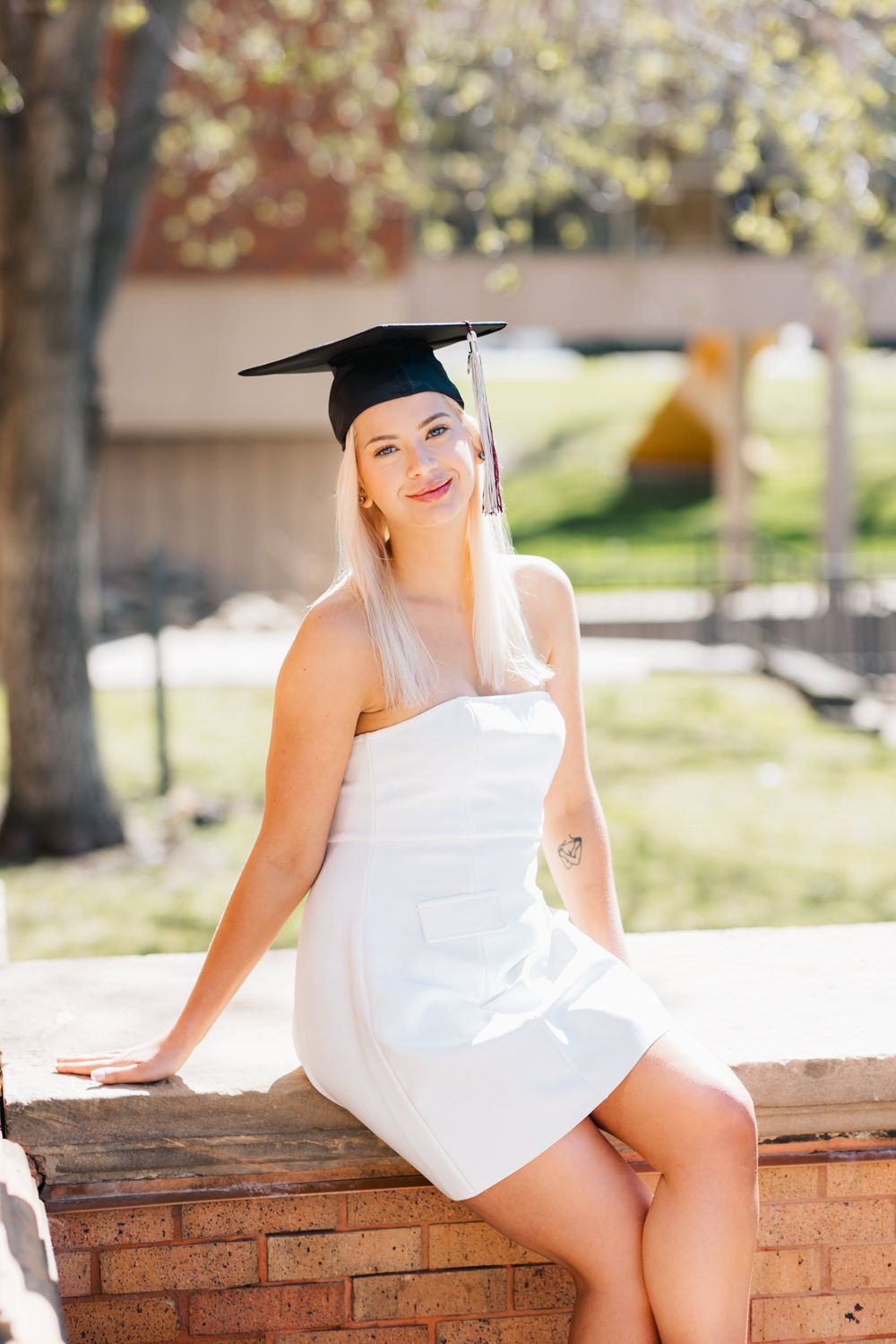 Graduate in white dress and cap sitting on brick ledge during outdoor photoshoot.