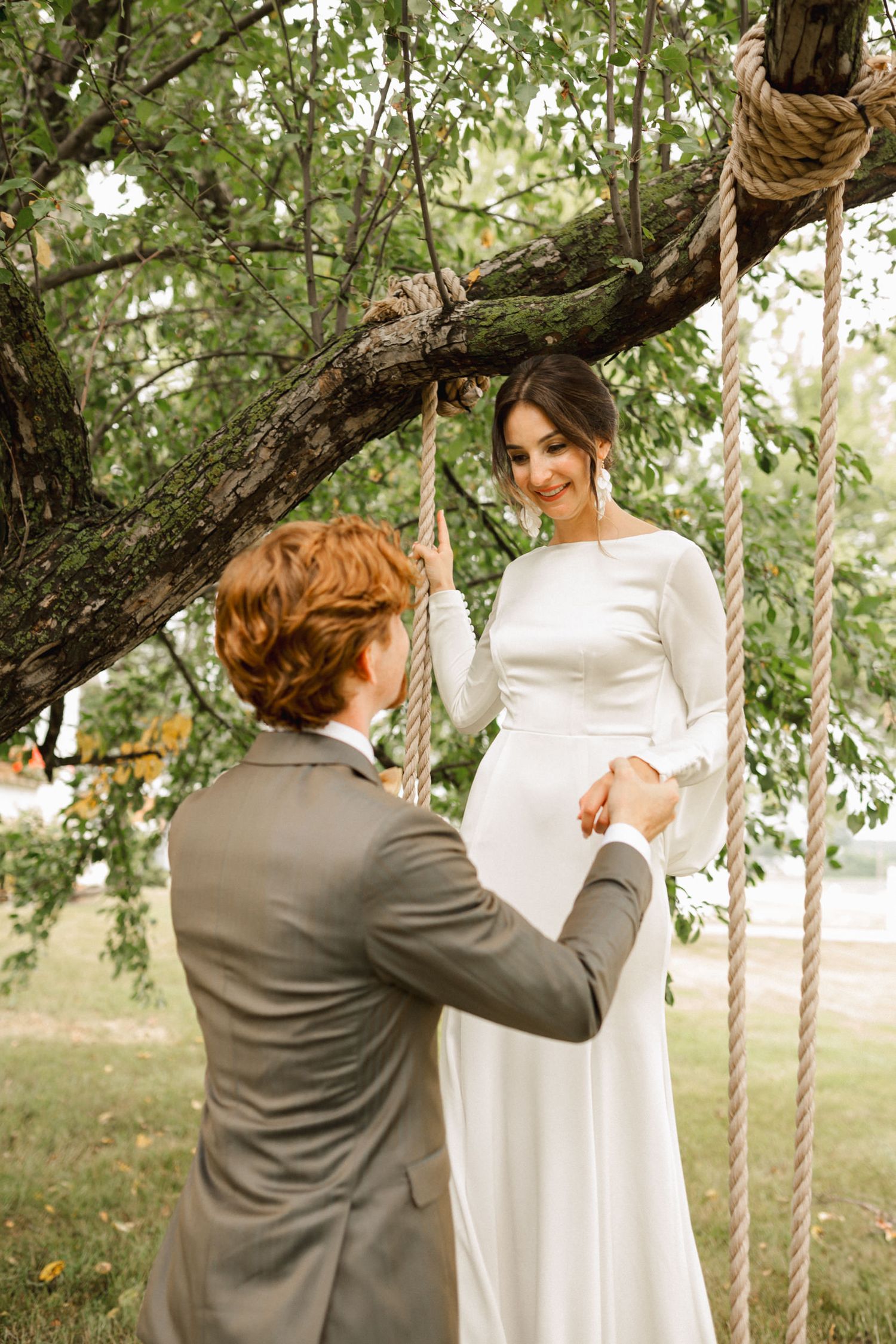 A romantic garden wedding scene with a couple at a tree swing in classic white dress and grey suit attire.