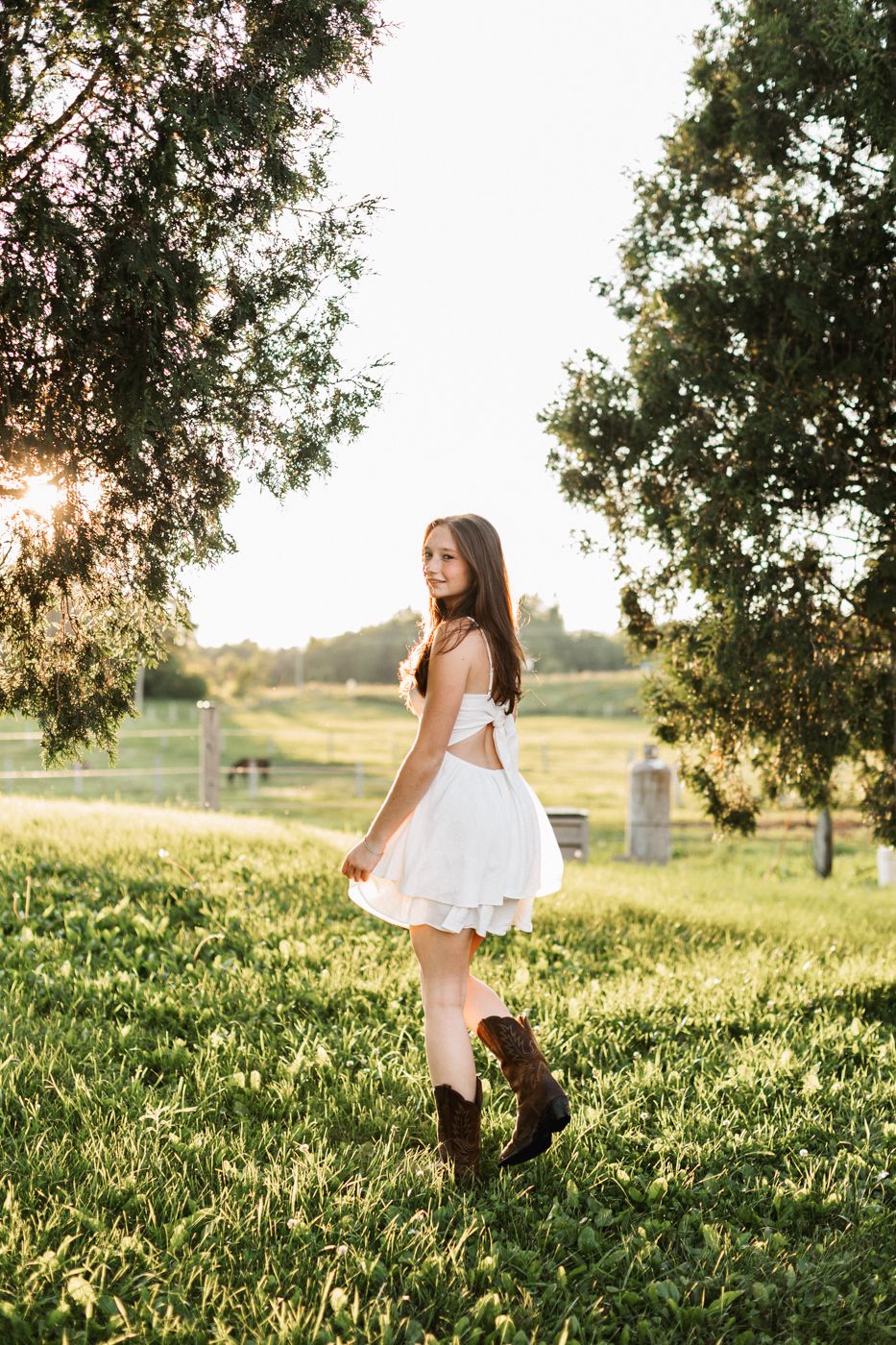 Figure in white dress and boots poses in golden sunlight with trees and fence in background.