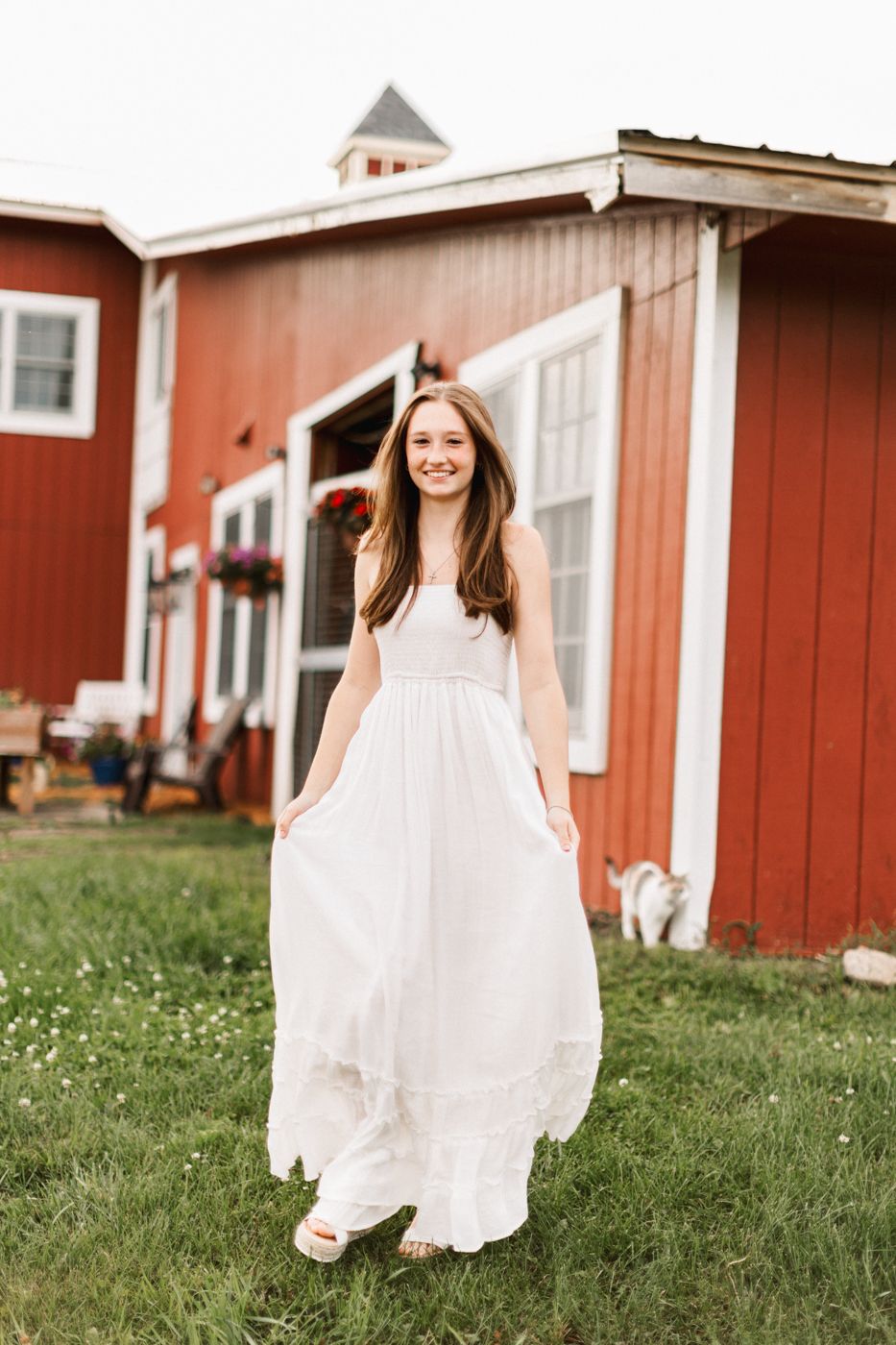 A full-length portrait in a flowing white dress outside a classic red barn.