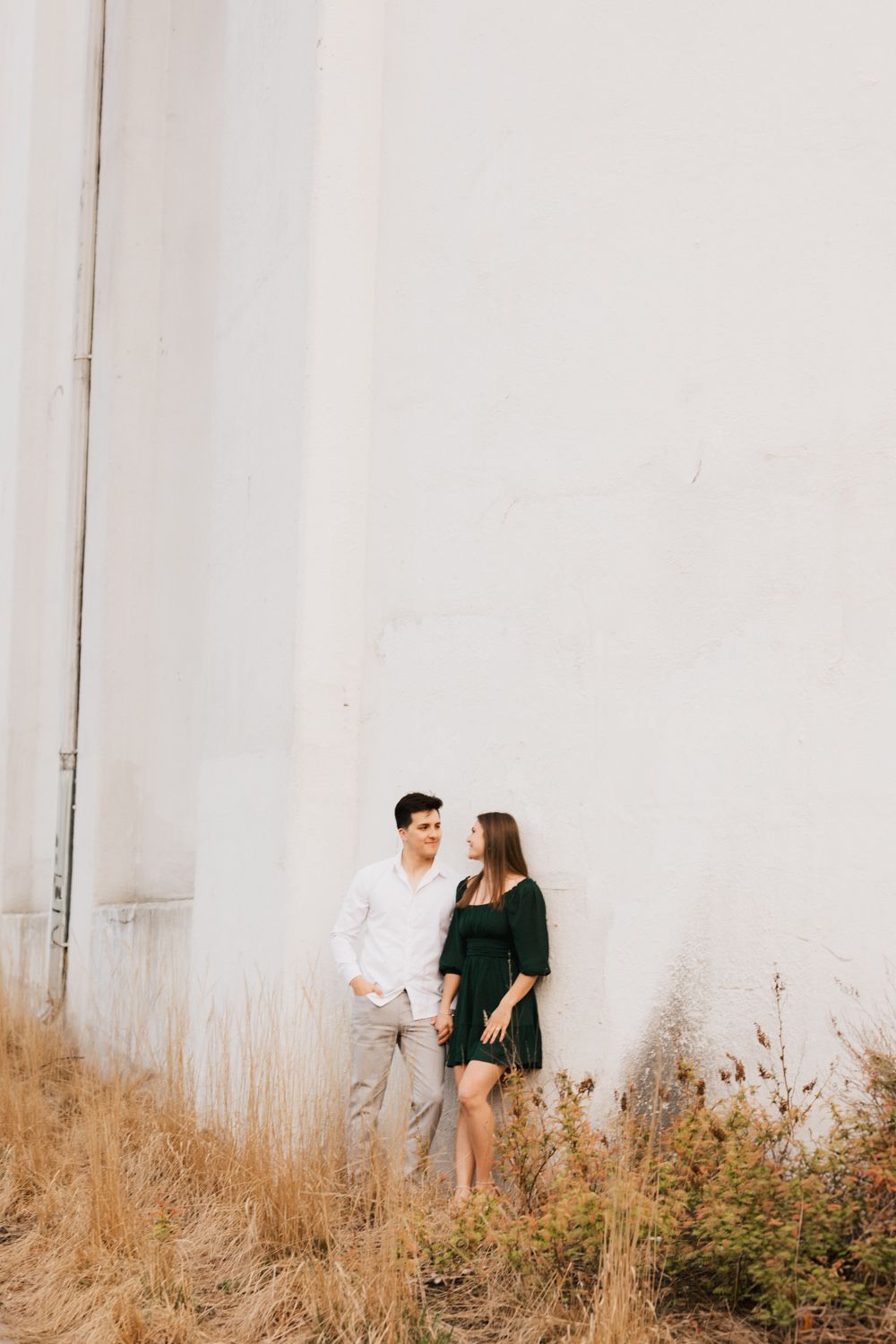 A couple standing together against a white wall surrounded by golden dry grass.