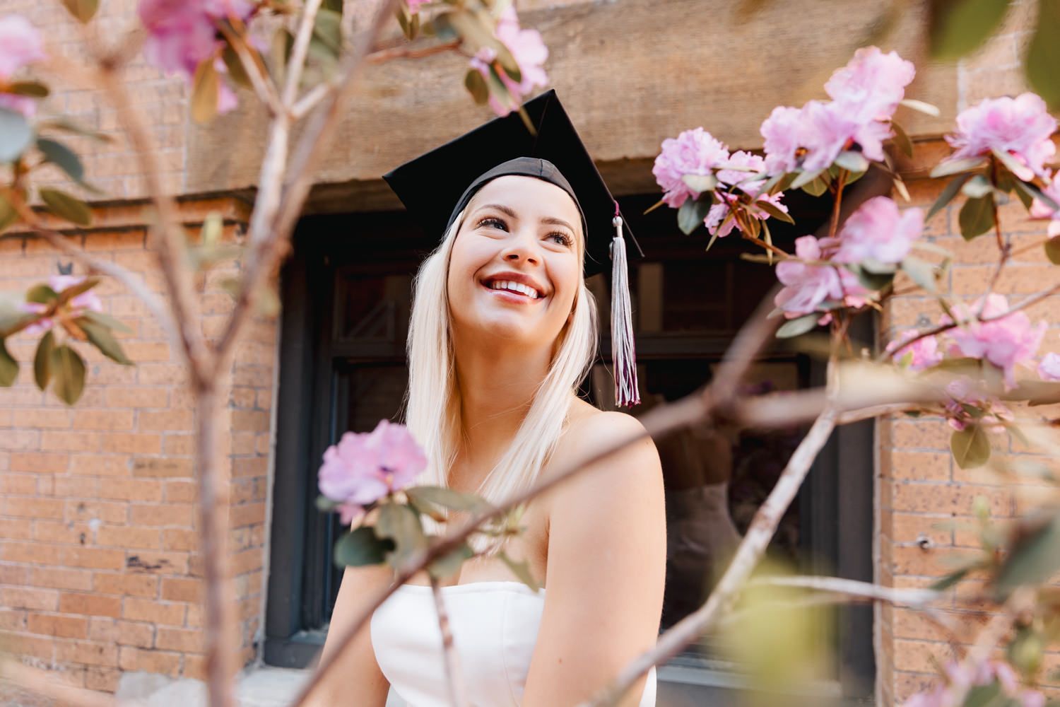 Graduate in black mortarboard smiles joyfully beneath blooming pink flowers.