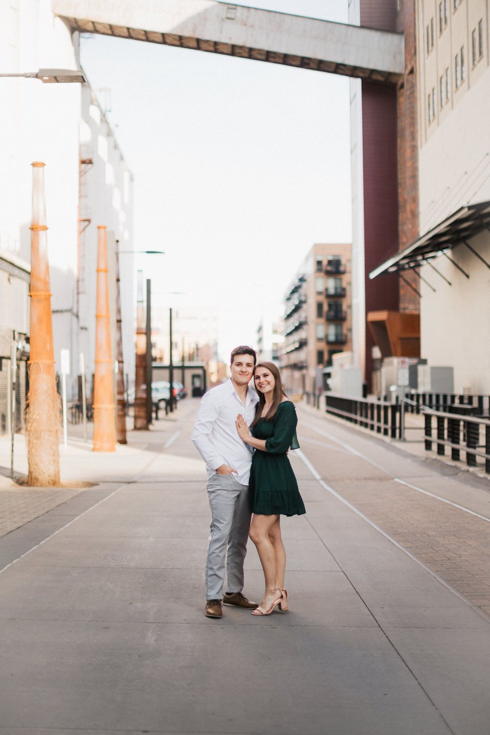 Couple posing together on urban sidewalk with woman in green dress and man in grey pants and white shirt.