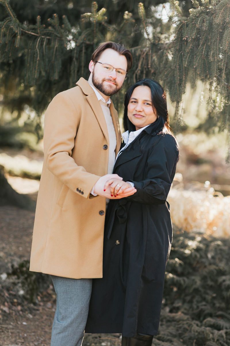 A couple poses together outdoors wearing elegant winter coats against a natural backdrop.