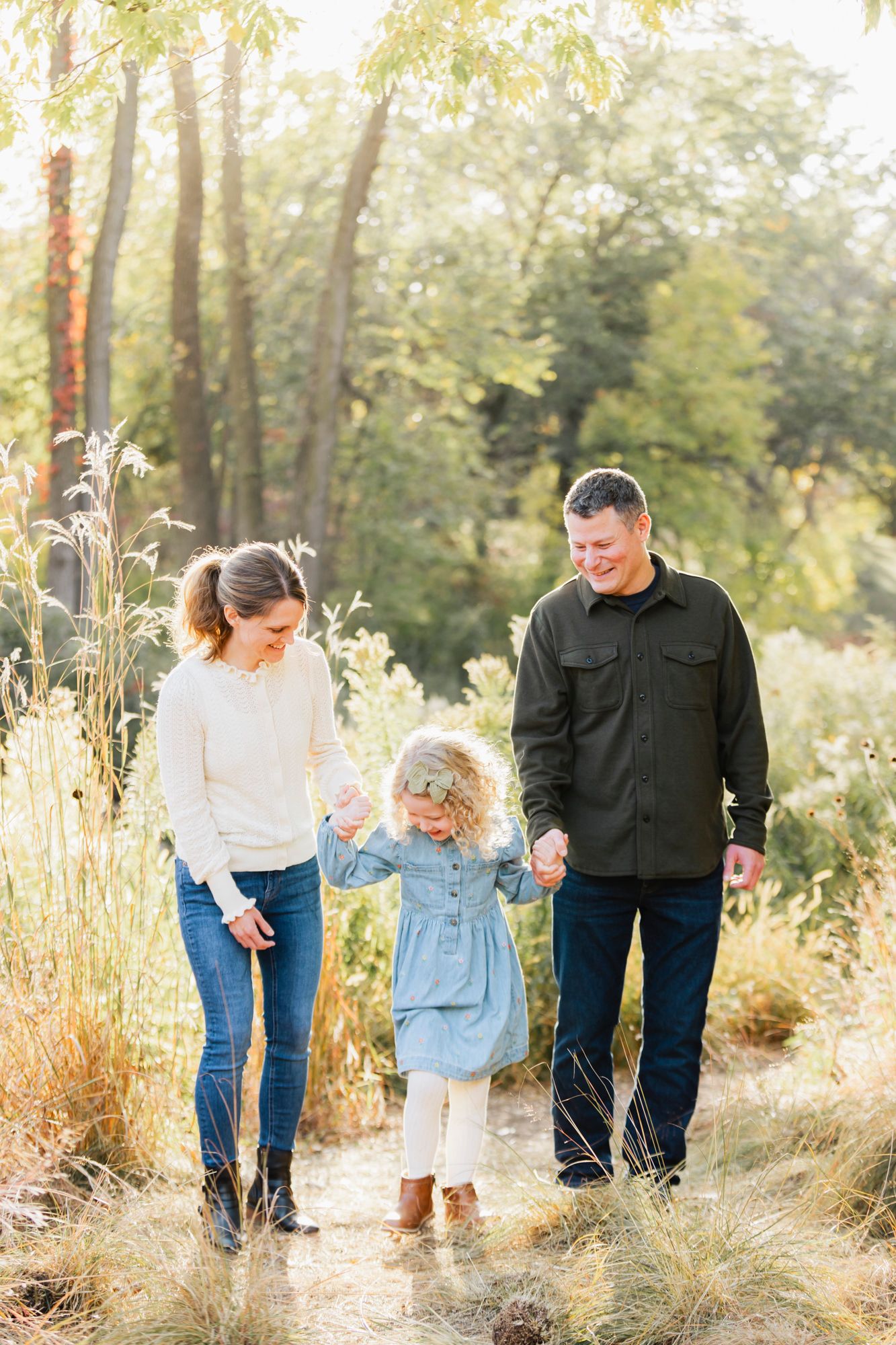 Family walks together on a nature trail surrounded by tall grass and trees in warm autumn light.