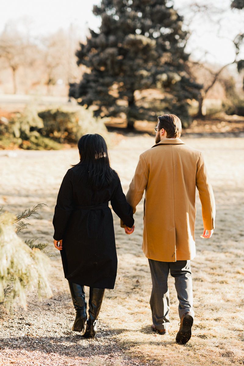 Couple in contrasting black and tan coats walking hand in hand on a nature path.