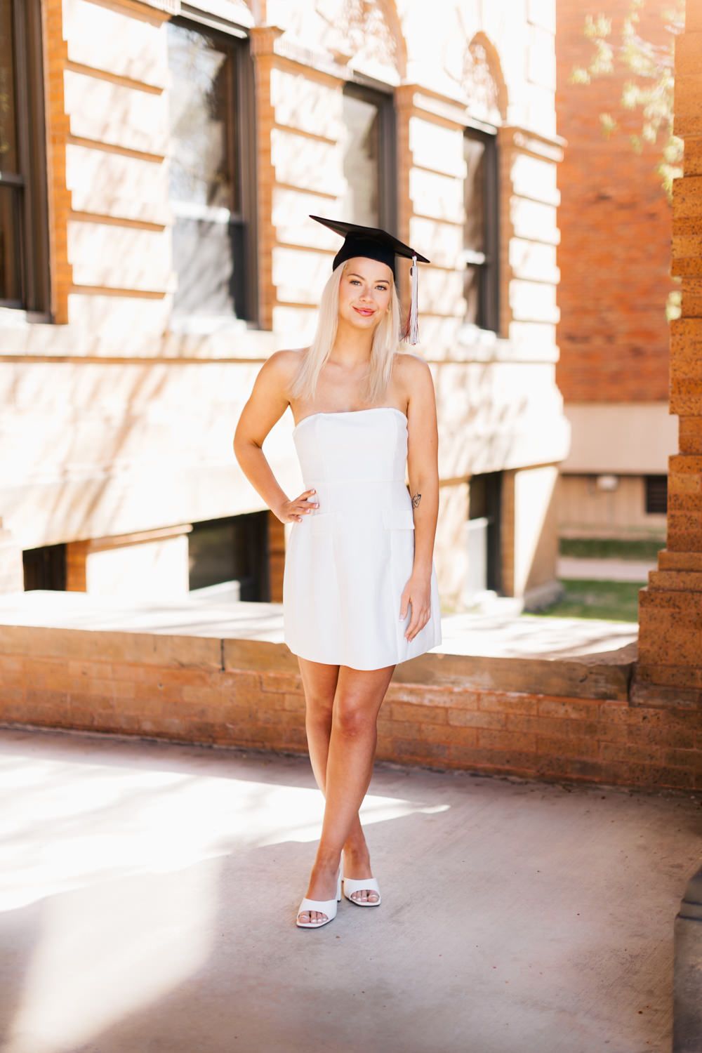 Graduate in short white dress and cap posing playfully by historic building columns.
