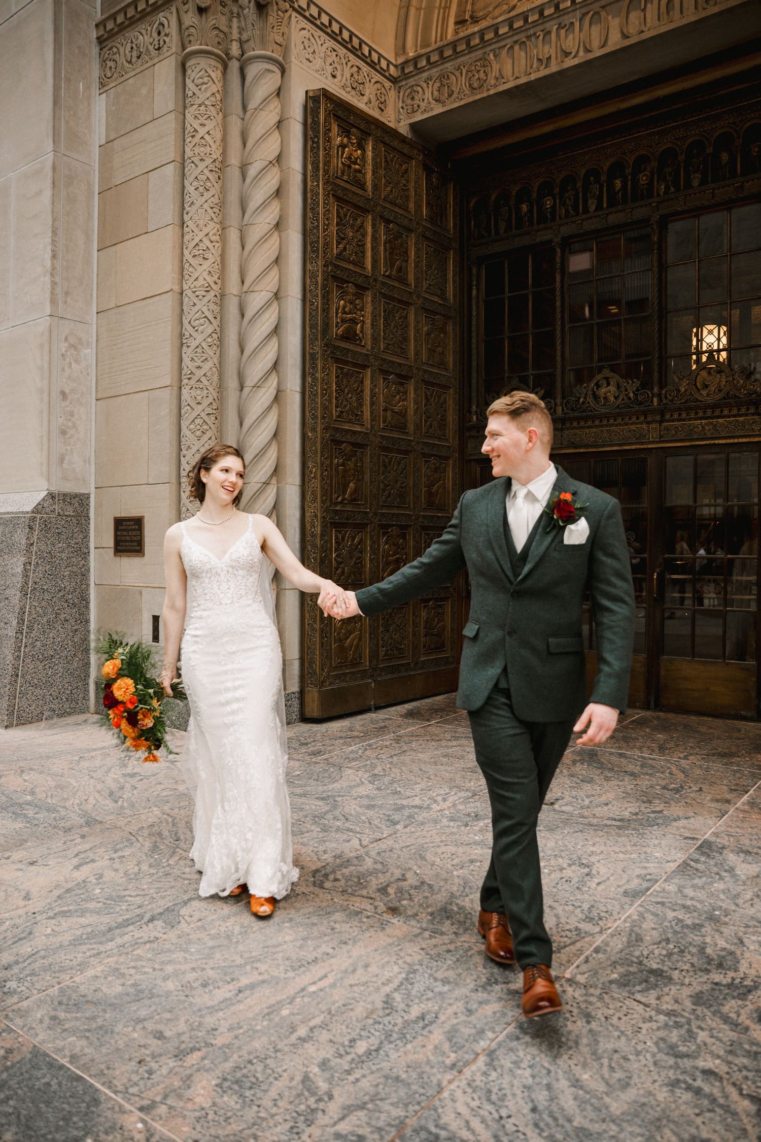 Elegant newlyweds walking hand in hand past ornate architectural details at their wedding venue.