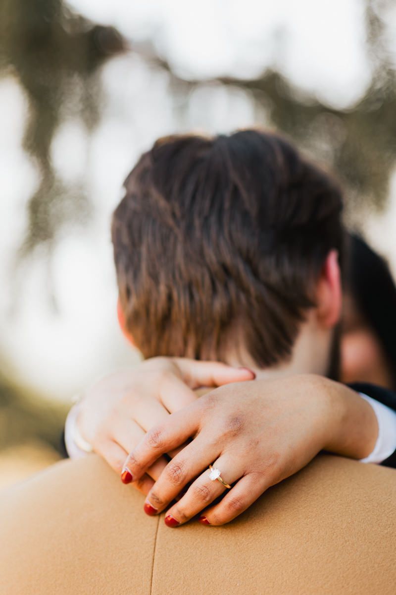 Close-up of hands with rings embracing on camel colored coat outdoors.