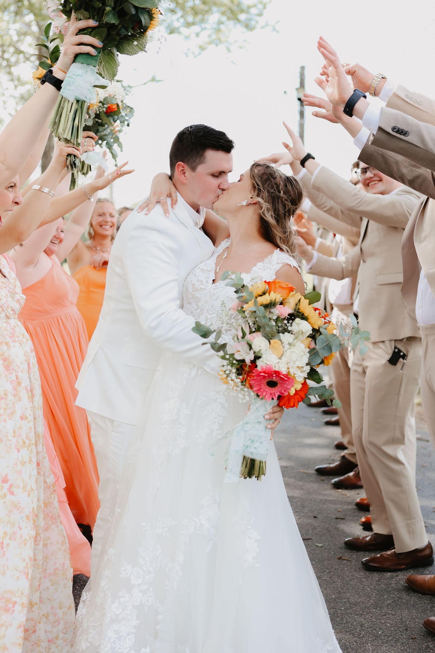 Bride and groom sharing a kiss under a celebratory walkway formed by their bridal party cheering, captured in bright, airy light that showca