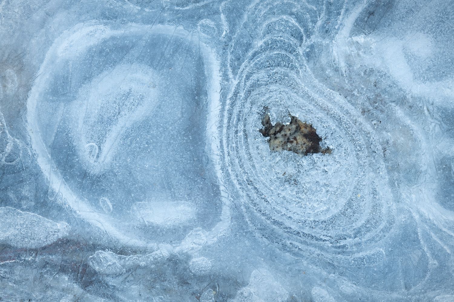 Abstract patterns in frozen ice surround a small dark object creating circular ripples.