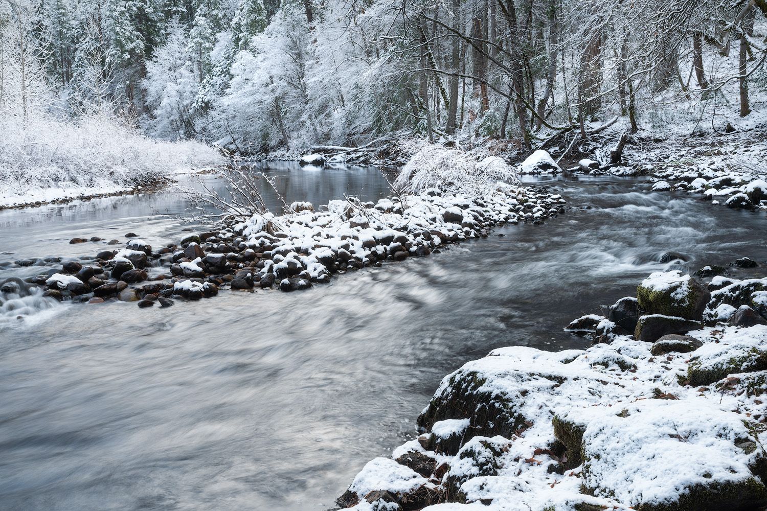 Snowy riverside landscape with ice-covered rocks and flowing water in winter.